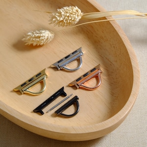 May include: Four different Apple Watch band adapters in silver, gold, rose gold, and black metal. The adapters are on a wooden surface.