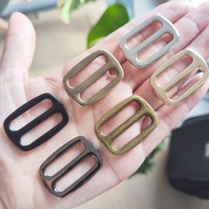 May include: A collection of metal slide buckles in various finishes: black, silver, gold, and antique brass. Each buckle has a rectangular shape with two parallel bars. These hardware components are likely used in bag making or strap adjustments.