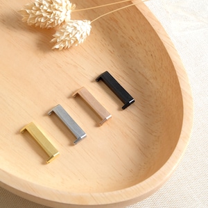 Puede incluir: Cuatro conectores de correa de reloj de metal en diferentes colores: dorado, plateado, dorado rosa y negro. Los conectores están dispuestos en una fila sobre una superficie de madera.