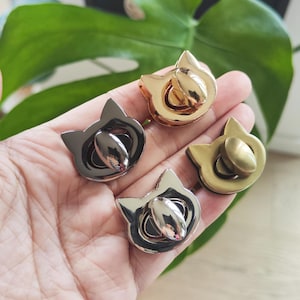 Könnte beinhalten: Vier metallene katzenförmige Geldbörsenverschlüsse in Gold-, Gunmetal- und Silbertönen. Die Verschlüsse haben eine abgerundete Mitte und ein Katzendesign. Die Verschlüsse werden in einer Hand mit grünem, blättrigem Hintergrund gehalten.