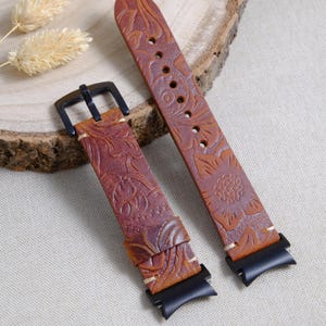 May include: Two-piece brown leather watch strap with a floral embossed design. The watch strap has a black buckle and keepers. The leather is a medium brown colour with darker brown accents. The watch strap is shown on a light-coloured surface.