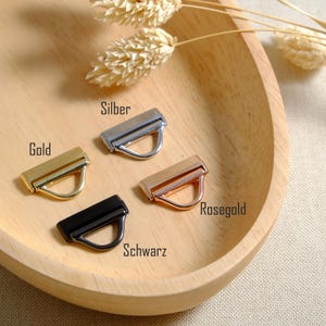 May include: Four metal purse hardware pieces in gold, silver, rose gold, and black, each with a rectangular bar and a D-ring. The hardware is displayed on a wooden tray with dried wheat stalks.
