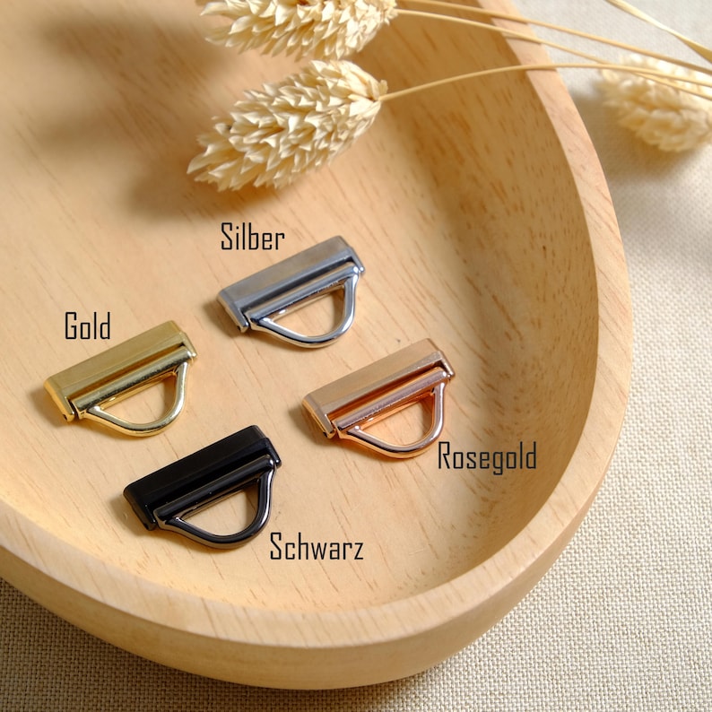 May include: Four metal purse hardware pieces in gold, silver, rose gold, and black, each with a rectangular bar and a D-ring. The hardware is displayed on a wooden tray with dried wheat stalks.