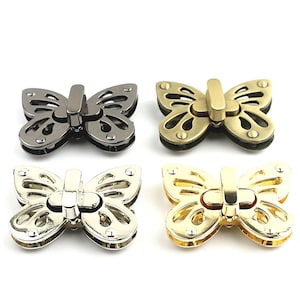 Könnte beinhalten: Vier Metall-Schmetterlingsverschlüsse in verschiedenen Ausführungen: Gunmetal, Antik-Messing, Silber und Gold. Jeder Verschluss hat eine Schmetterlingsform mit filigranen Ausschnitten und einem zentralen Verriegelungsmechanismus. Geeignet für Taschen und Accessoires.