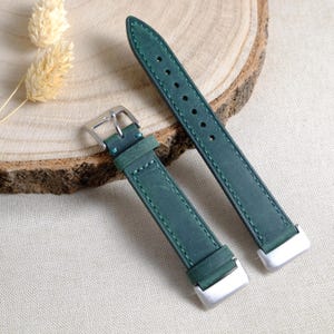 May include: Two-piece, dark green leather watch strap with silver-coloured metal hardware. The strap is stitched with a lighter green thread. The watch band is displayed on a wooden surface.