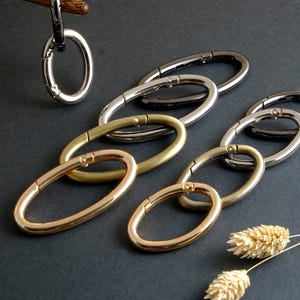 May include: Oval-shaped metal clasps in various finishes: silver, black, gold, and brass. The clasps are arranged on a dark surface, with one clasp suspended from a wooden element. The image showcases the different color options and the clasp's design.