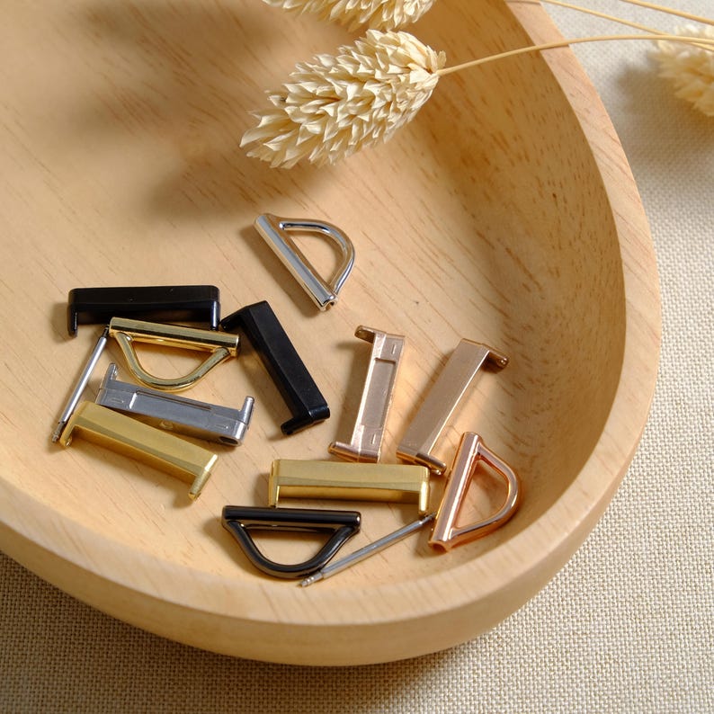 May include: Assortment of metal D-ring and slider buckles in gold, silver, and black finishes. The buckles are arranged on a wooden surface.