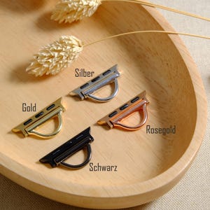May include: Four watch band adapters in gold, silver, rose gold, and black, displayed on a wooden tray. The adapters have a curved, D-shaped loop for attaching to a watch band. The words "Gold", "Silber", "Rosegold", and "Schwarz" are visible.