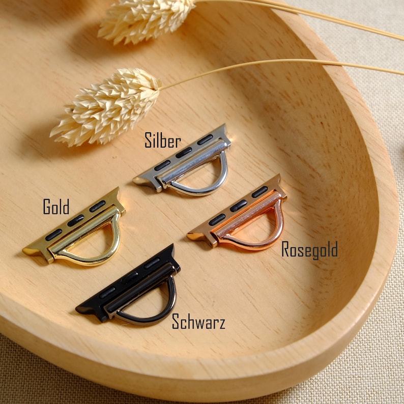 May include: Four watch band adapters in gold, silver, rose gold, and black, displayed on a wooden tray. The adapters have a curved, D-shaped loop for attaching to a watch band. The words "Gold", "Silber", "Rosegold", and "Schwarz" are visible.