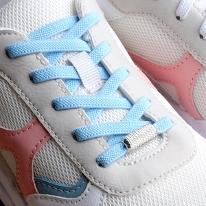 Può includere: Primo piano di un paio di scarpe da ginnastica bianche con lacci azzurri. Le scarpe da ginnastica hanno dettagli rosa e blu, un tessuto a rete e un fermalacci in metallo argentato. I lacci sono strutturati.