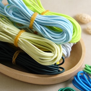 May include: A close-up of a variety of colorful elastic cords in a wooden bowl. The cords are in shades of green, blue, yellow, and black.