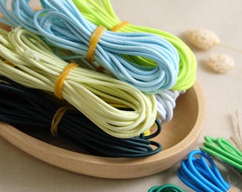 Può includere: Primo piano di una varietà di cordoni elastici colorati in una ciotola di legno. I cordoni sono in tonalità di verde, blu, giallo e nero.