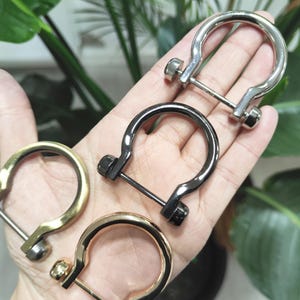 May include: Four metal D-ring buckles in silver, black, gold, and brass. Each has a U-shaped ring with a screw-in bar. The buckles are held in a hand, with green foliage in the background. Measurements are in cm.