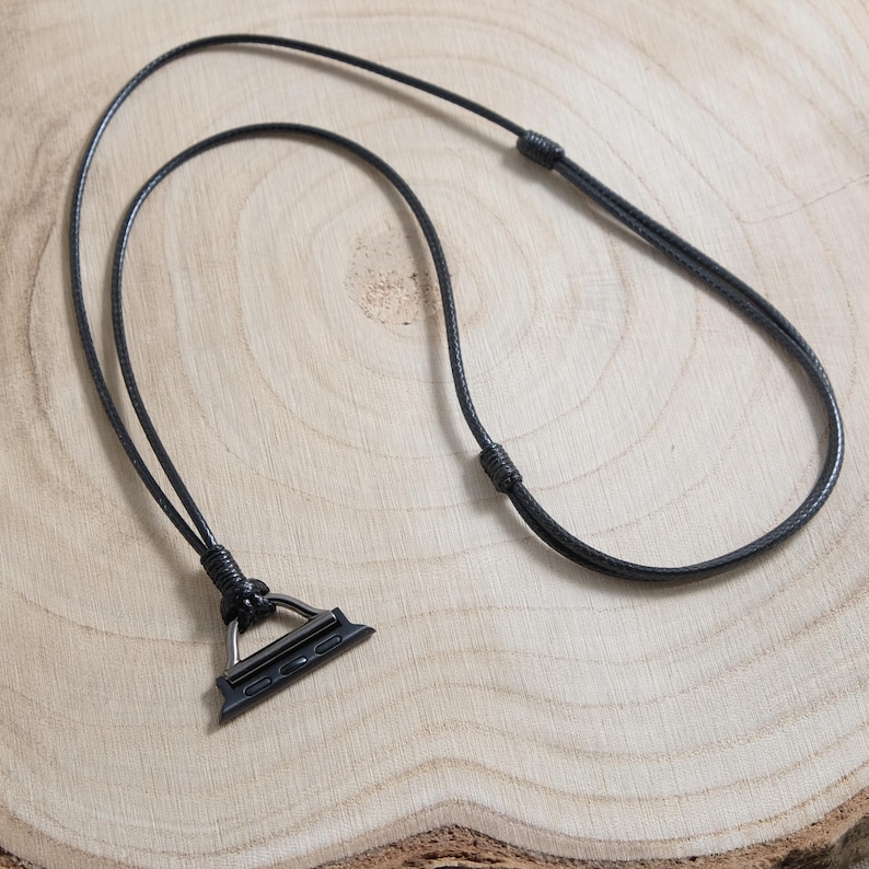 May include: A black leather Apple Watch lanyard with a black metal connector. The lanyard is made of a thin, braided cord and is designed to be worn around the neck. The watch connector is approximately 2.5 cm wide.