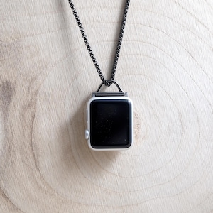 May include: A rectangular smartwatch with a black screen and a white casing, suspended from a dark metallic box chain necklace. The device features a small silver side button and a top-mounted metal loop attachment for the chain to pass through.
