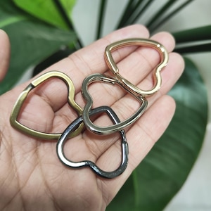 Puede incluir: Un conjunto de llaveros metálicos en forma de corazón en varios acabados: dorado, latón, plateado y pavonado. Los llaveros están entrelazados, mostrando sus variaciones de diseño y color. El diseño en forma de corazón es una característica destacada.