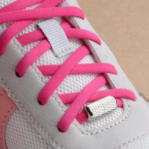 Könnte beinhalten: Nahaufnahme eines weißen und rosa Sportschuhs mit leuchtend pinken Schnürsenkeln. Die Schnürsenkel sind durch weiße Ösen und eine graue Stoffschlaufe gefädelt. Ein silbernes Metall-Aglet sichert die Schnürsenkel.