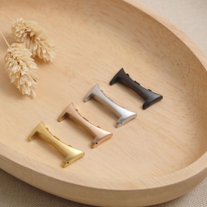 Könnte beinhalten: Vier Uhrenarmband-Adapter aus Metall, die in einer diagonalen Reihe angeordnet sind und in Gold, Roségold, Silber und Schwarz ausgeführt sind. Jeder Adapter hat eine geschwungene Kante und kleine Befestigungspunkte für das Uhrengehäuse.