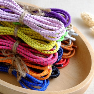 Puede incluir: Conjuntos de coloridos cordones elásticos en varios tonos, incluyendo rosa, amarillo, morado y azul, atados con hilo. Los cordones tienen un patrón moteado y están dispuestos en un cuenco de madera.