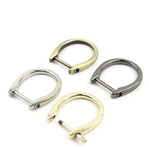 Könnte beinhalten: Vier D-Ringe aus Metall in verschiedenen Ausführungen: Gold, Silber, Antikmessing und Schwarz. Die Ringe sind alle gleich groß und haben die gleiche Form und ein schlichtes, elegantes Design.