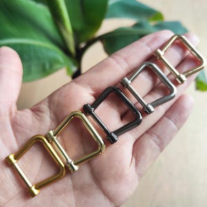 Puede incluir: Una mano muestra cinco hebillas cuadradas de metal en acabados dorado, plateado y negro. Las hebillas tienen forma rectangular con un detalle de tornillo. Hojas verdes en el fondo.
