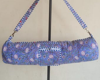 May include: A cylindrical yoga mat bag featuring a blue fabric with a repeating floral pattern in white and red. The bag includes a matching adjustable shoulder strap with a blue and white striped border and a central rectangular pocket with a zipper.
