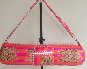 May include: A cylindrical yoga mat bag featuring a vibrant pattern of orange and pink vertical stripes with repeating tiger illustrations. The bag includes a white zippered pocket on the front, a matching adjustable shoulder strap, and white trim.