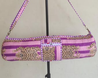 May include: A cylindrical yoga mat bag featuring a repeating tiger pattern over a pink and purple striped background. The bag includes a matching adjustable shoulder strap, a central zippered pocket, and white and purple checkered trim detailing.