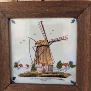 Può includere: Piastrella in ceramica incorniciata con una scena di mulino a vento. L'opera raffigura un tradizionale mulino a vento olandese con tetto di paglia, su sfondo azzurro. La cornice in legno aggiunge un tocco rustico.