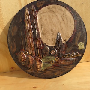 Op de afbeelding: Een rond keramisch wandkunstwerk met een bruin, groen en gouden kleurenschema. Het stuk toont een gestileerd landschap met bomen en bladeren. De tekst "BUSCH" en "Art & Handwerk" is zichtbaar in de rechterbenedenhoek.