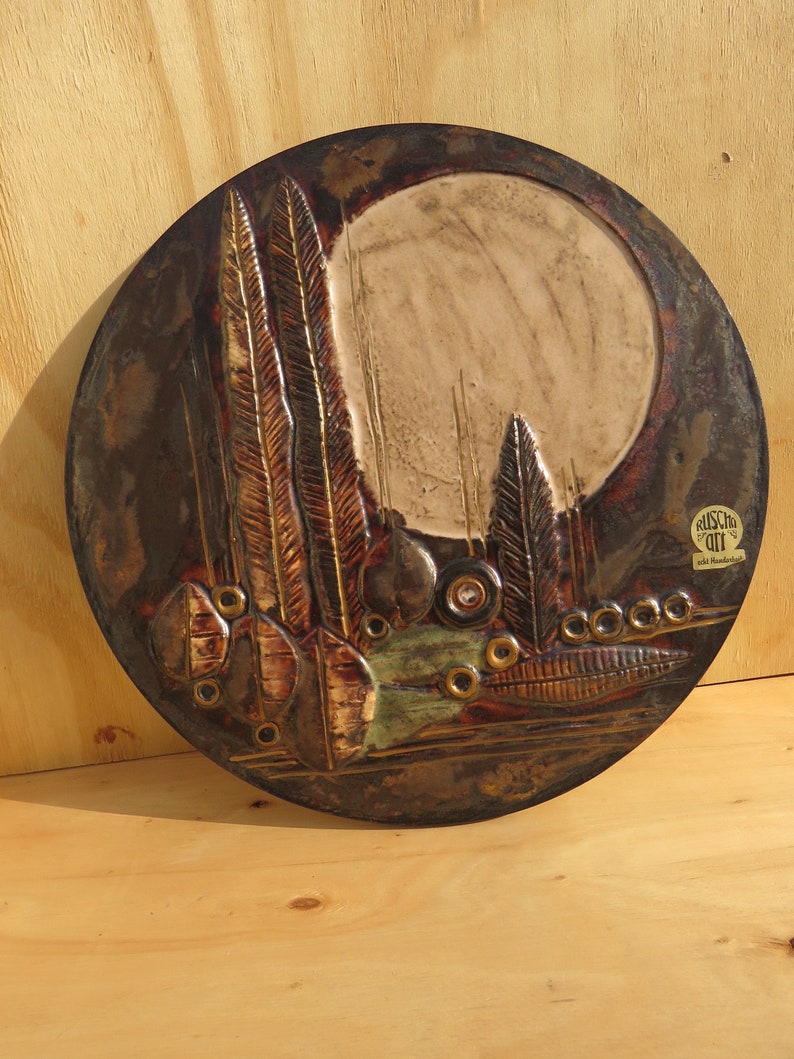 Op de afbeelding: Een rond keramisch wandkunstwerk met een bruin, groen en gouden kleurenschema. Het stuk toont een gestileerd landschap met bomen en bladeren. De tekst "BUSCH" en "Art & Handwerk" is zichtbaar in de rechterbenedenhoek.
