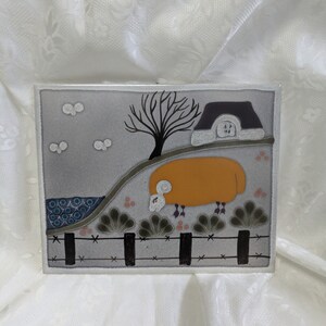 Może przedstawiać: Płytka ceramiczna z fantazyjną ilustracją owcy stojącej na wzgórzu z małym domkiem w tle. Owca jest pomarańczowa z białą twarzą i czarnymi kopytami. Domek jest szary z fioletowym dachem i białym oknem. Płytka jest ozdobiona czarnym płotem i zielonymi krzewami.