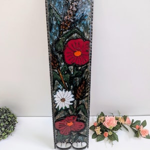 Könnte beinhalten: Ein langes, rechteckiges Wanddekorationsstück mit floralem Design. Das Stück hat einen Metallrahmen und einen Keramikflieseneinsatz mit roten, weißen und grünen Blumen auf blauem Hintergrund. Das Stück ist etwa 61 cm hoch.