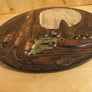 Op de afbeelding: Een ronde, bruine keramische wanddecoratie met een reli&euml;fdesign van bomen, bladeren en cirkels. De wanddecoratie heeft een witte achtergrond en een bruine rand. De tekst "Rustic Art" is zichtbaar op een label dat aan de wanddecoratie is bevestigd.
