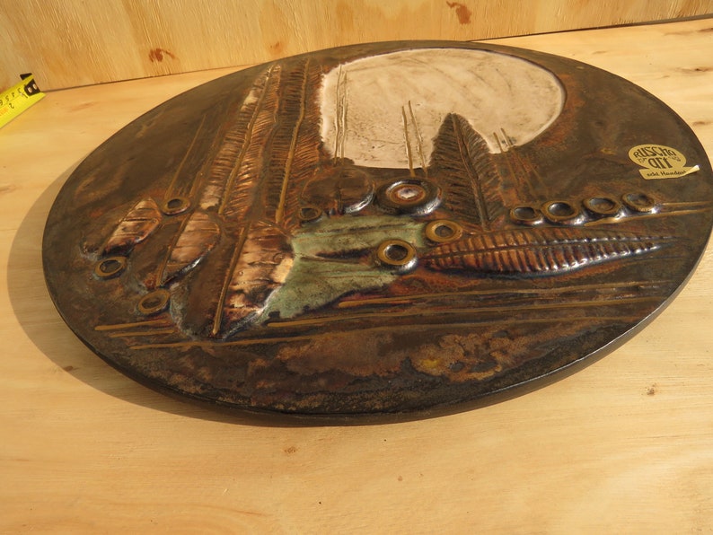 Op de afbeelding: Een ronde, bruine keramische wanddecoratie met een reli&euml;fdesign van bomen, bladeren en cirkels. De wanddecoratie heeft een witte achtergrond en een bruine rand. De tekst "Rustic Art" is zichtbaar op een label dat aan de wanddecoratie is bevestigd.