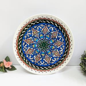 Puede incluir: Un plato de cerámica decorativo con un diseño floral colorido y detallado. El plato presenta un patrón central en forma de estrella en verde, rodeado de motivos florales azules, rojos y amarillos. El borde es blanco con un borde festoneado y un borde marrón y verde.