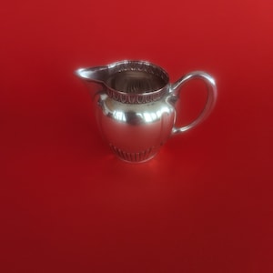 Może przedstawiać: Srebrny dzbanek do śmietanki z ozdobnym rantem i zaokrąglonym korpusem. Dzbanek stoi na czerwonej powierzchni.