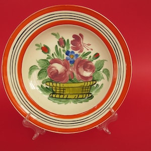 Op de afbeelding: Een wit keramiek bord met een handgeschilderd bloemenmotief. Het bord heeft een brede oranje rand met zwarte strepen. Het bloemenmotief toont roze rozen, blauwe bloemen en groene bladeren in een mand.