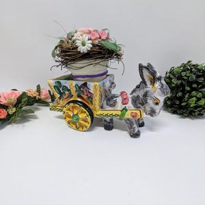 Può includere: Statuina in ceramica di un asino che tira un carretto decorativo pieno di un nido di fiori rosa e bianchi. Il carretto è dipinto con dettagli gialli, verdi e arancioni. L'asino è grigio e bianco.