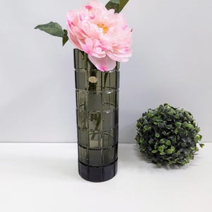 Op de afbeelding: Een donkergrijze vaas met rechthoekig patroon houdt een grote roze pioenroos met groene bladeren vast. Rechts van de vaas staat een kleine, ronde, groene topiary. De vaas staat op een wit oppervlak.