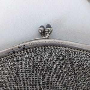 May include: A silver mesh purse with a clasp closure and the inscription "Marcelle Kuno" on the front.