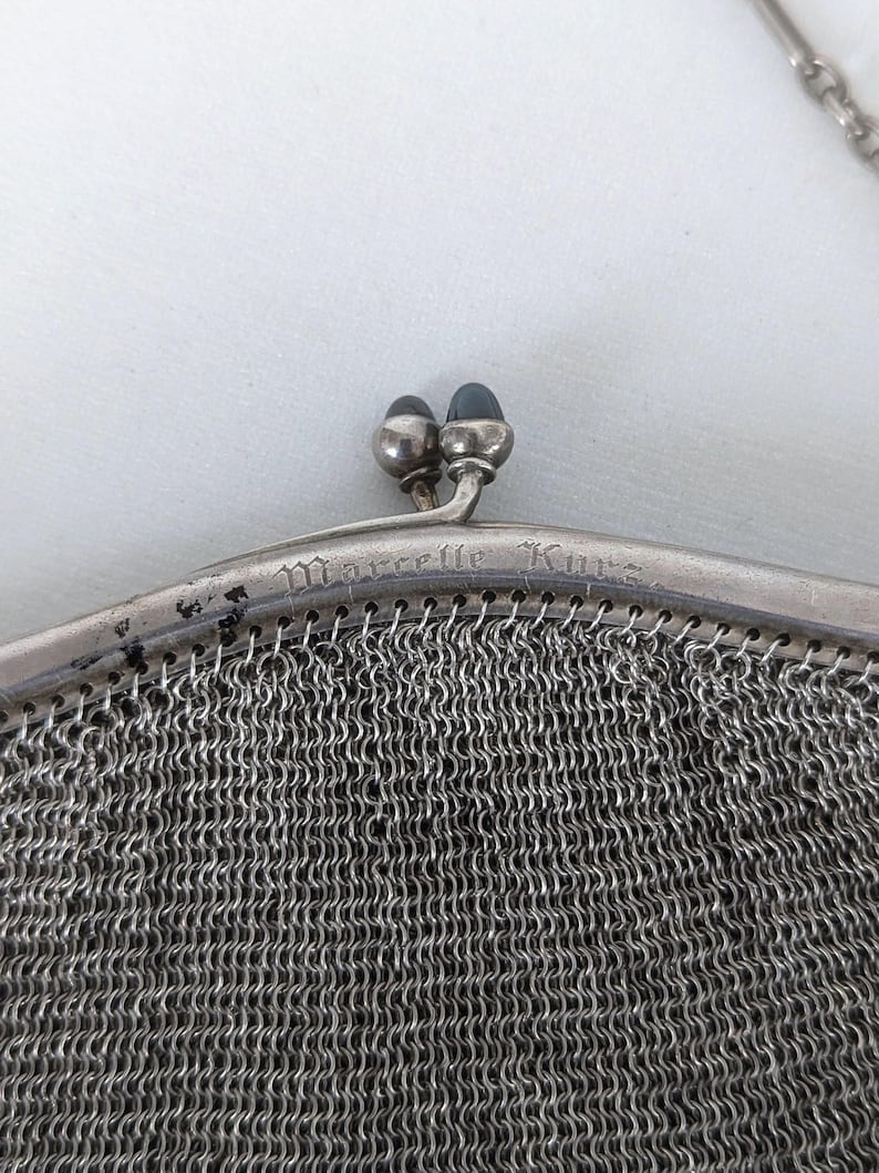 May include: A silver mesh purse with a clasp closure and the inscription "Marcelle Kuno" on the front.