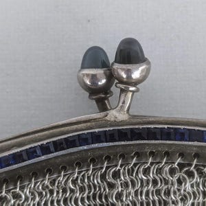 May include: A silver mesh purse with a top closure featuring two dark blue cabochon stones. The purse is decorated with a row of smaller blue stones around the top edge.