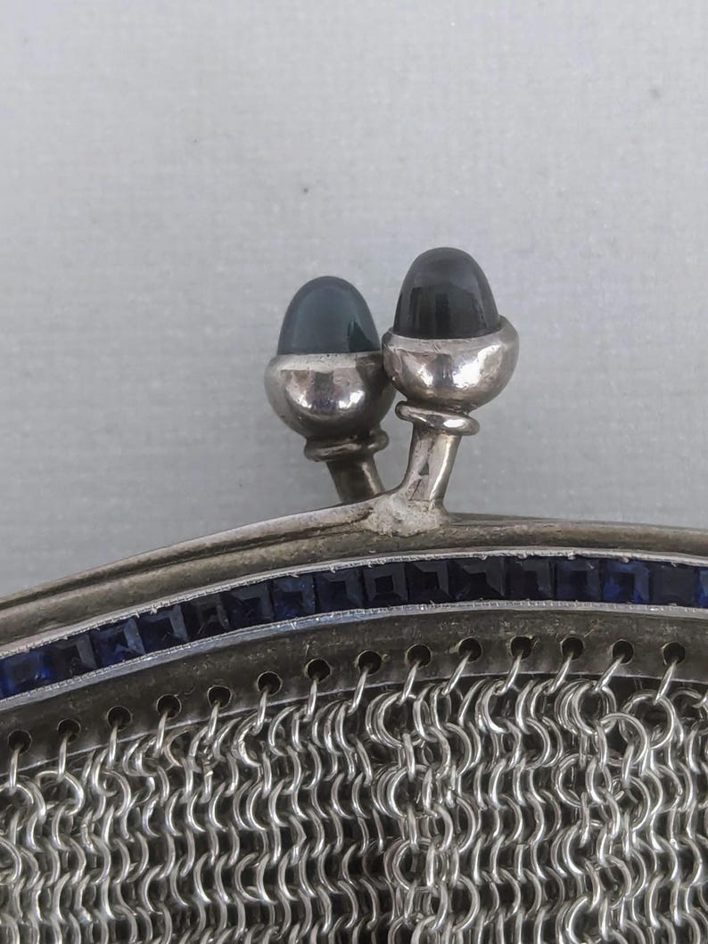 May include: A silver mesh purse with a top closure featuring two dark blue cabochon stones. The purse is decorated with a row of smaller blue stones around the top edge.