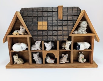 May include: A wooden house-shaped shelf unit with multiple compartments, each housing a small ceramic cat figurine. The roof is dark brown, and the frame is light brown. The cats are in various poses and colors.