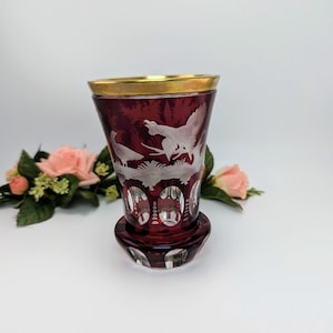 May include: A ruby red glass with a gold rim and a matching base. The glass features etched white bird designs. The glass is likely a vintage item, possibly for serving beverages. The glass is approximately 10 cm tall.
