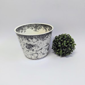 May include: A white ceramic planter with a black floral pattern. The planter is empty and sitting on a white surface. A small green bush is sitting to the right of the planter.