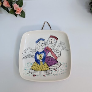 Peut inclure: Assiette murale carrée en céramique avec une illustration peinte à la main de deux personnages en robes et coiffes colorées. Le fond représente un paysage avec une ville et des montagnes. Une corde marron est attachée pour l'accrocher.