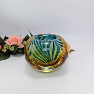 Op de afbeelding: Een ronde glazen kom met een wervelontwerp in tinten geel, groen en blauw. De kom heeft een brede opening en een afgeronde basis. Het glas is transparant en laat licht door. De kom wordt waarschijnlijk gebruikt voor decoratieve doeleinden.