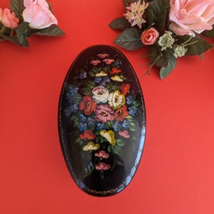 May include: An oval-shaped black lacquered box with a floral design painted on the lid. The flowers are red, pink, white, blue, and yellow. The box has a gold trim around the edge.